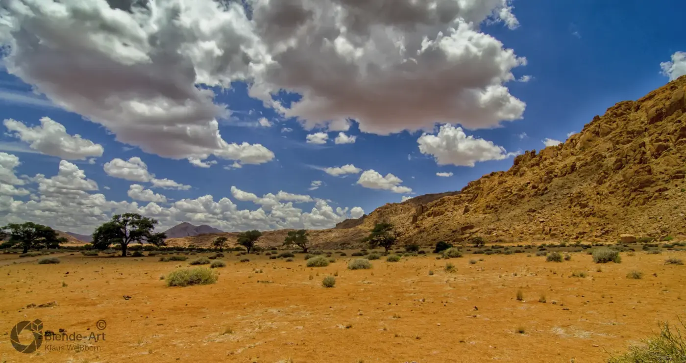 Trockene Landschaft bei der Spitzkoppe in Namibia mit markanter Felswand, Bäumen und blauem Himmel voller Quellwolken