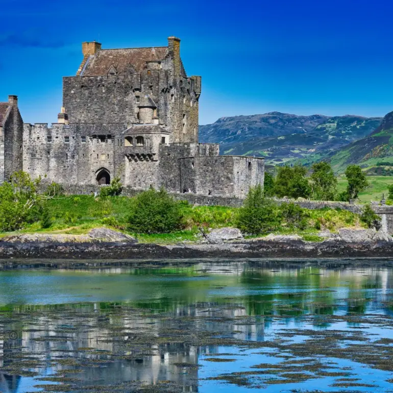 Eilean Donan Castle mit Spiegelung im Wasser vor grünen Highlands in Schottland bei Sonnenschein.