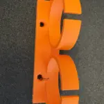 Rückansicht einer orangefarbenen 3D-gedruckten Dremel Doppelhalterung aus PETG mit zwei Senkbohrungen zur Wandbefestigung, auf dunkler Oberfläche fotografiert.
