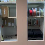 Schrank mit Glasschiebetüren und montierten Filamentrollenhaltern zur übersichtlichen Lagerung von 3D-Druck Filamentrollen