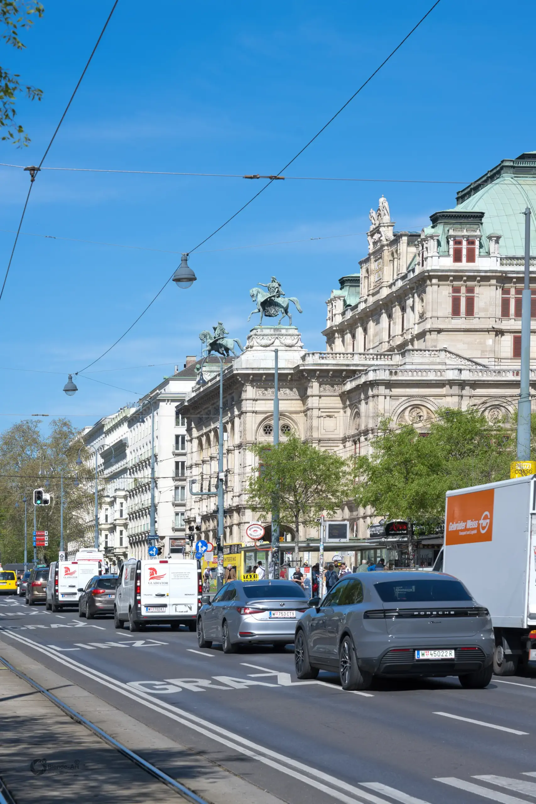 Blick entlang des Wiener Opernrings auf die historische Fassade der Staatsoper mit fließendem Autoverkehr und LKWs bei Tageslicht.