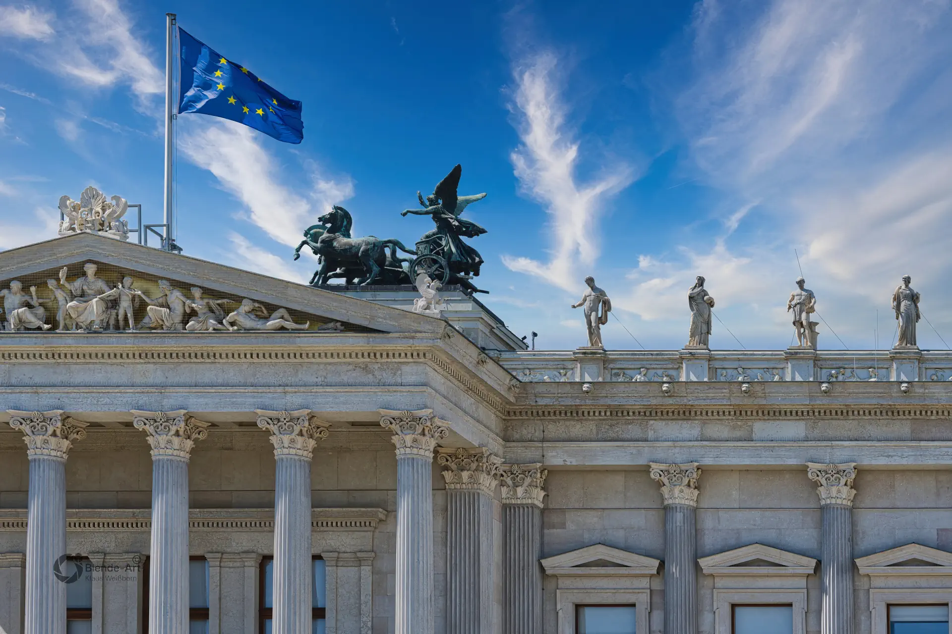 Blick auf das Dach des Wiener Parlaments mit einer antiken Quadriga-Skulptur und der wehenden EU-Flagge vor blauem Himmel.