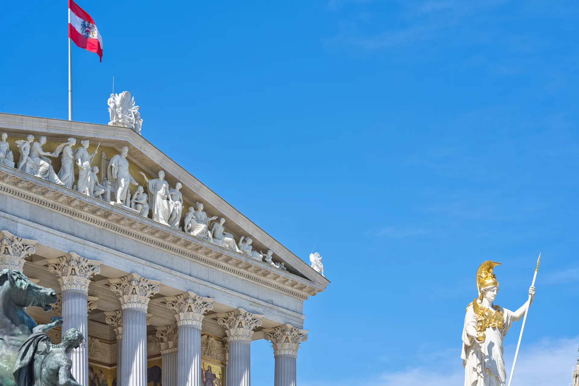 Detailaufnahme des Wiener Parlamentsgebäudes mit der Pallas-Athene-Statue und dem figurenreichen Giebelrelief vor strahlend blauem Himmel.