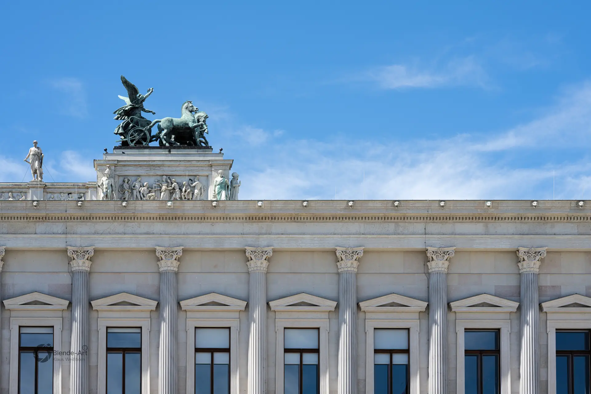 Detailaufnahme der Attika des Wiener Parlaments mit einer bronzenen Quadriga und antiken Statuen vor blauem Himmel.