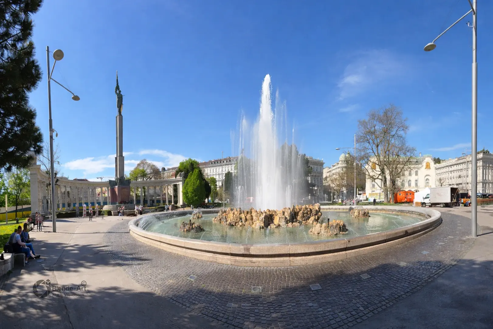 Weitwinkel-Panorama des Wiener Schwarzenbergplatzes mit dem aktiven Hochstrahlbrunnen und dem sowjetischen Heldendenkmal unter blauem Himmel.
