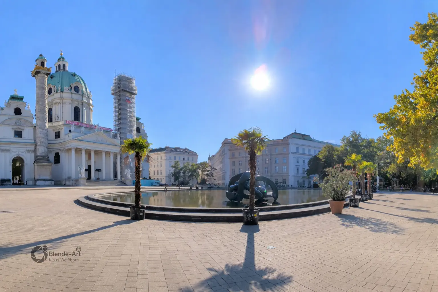 Panorama-Ansicht des Wiener Karlsplatzes mit der barocken Karlskirche, dem vorgelagerten Brunnen und herbstlichen Bäumen unter sonnig blauem Himmel.