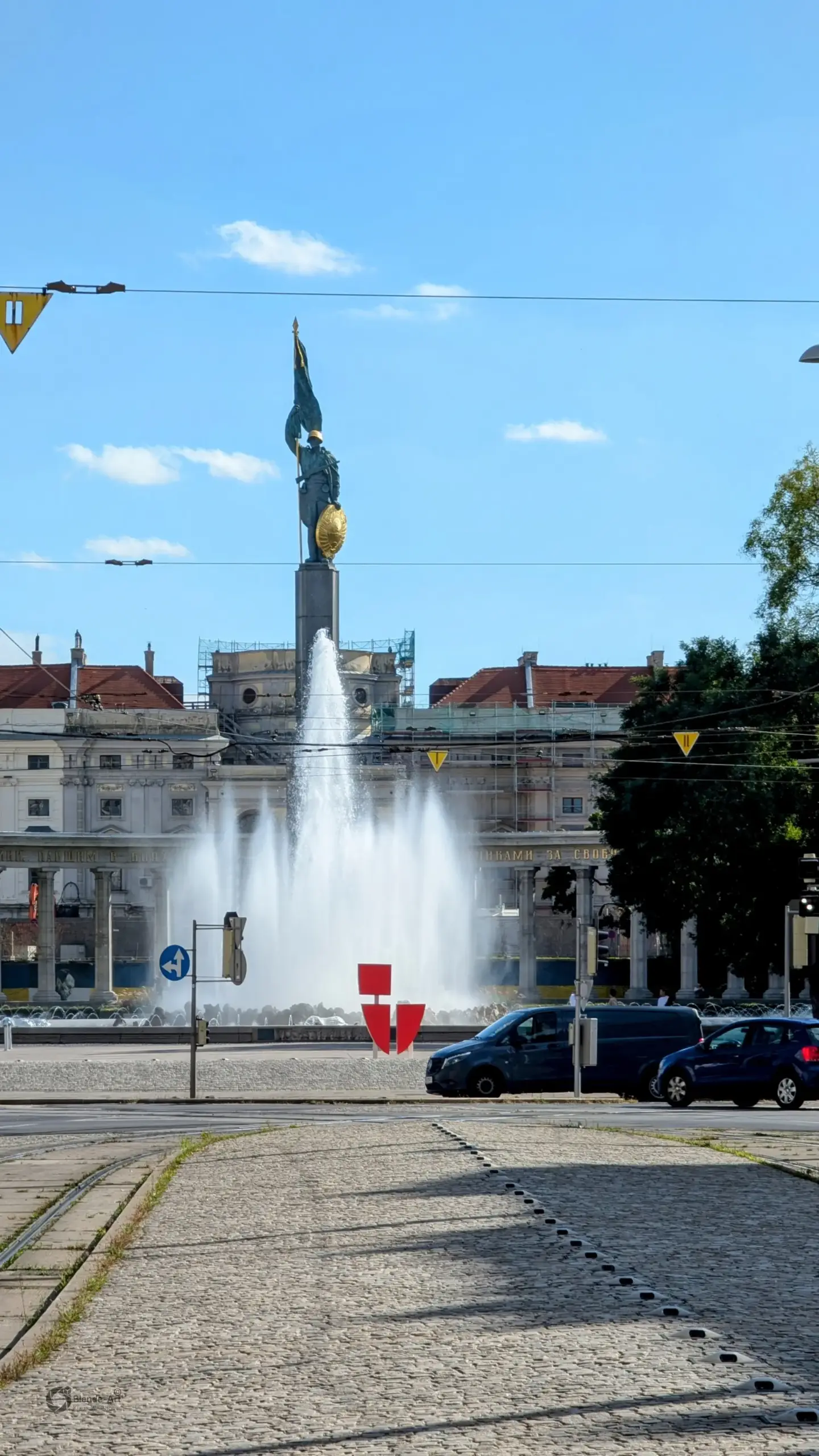 Blick über das Kopfsteinpflaster am Wiener Schwarzenbergplatz auf den aktiven Hochstrahlbrunnen und das sowjetische Heldendenkmal unter blauem Himmel.