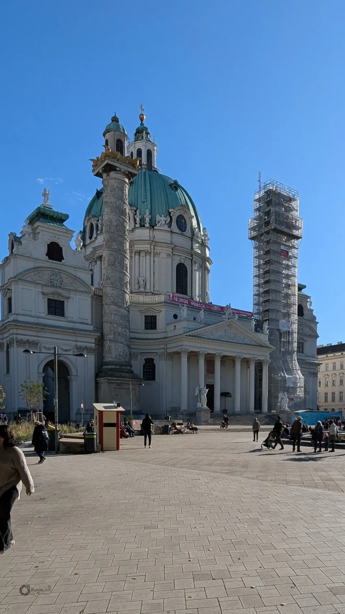 Nahaufnahme der barocken Karlskirche in Wien mit einer eingerüsteten Reliefsäule und Passanten auf dem gepflasterten Karlsplatz.