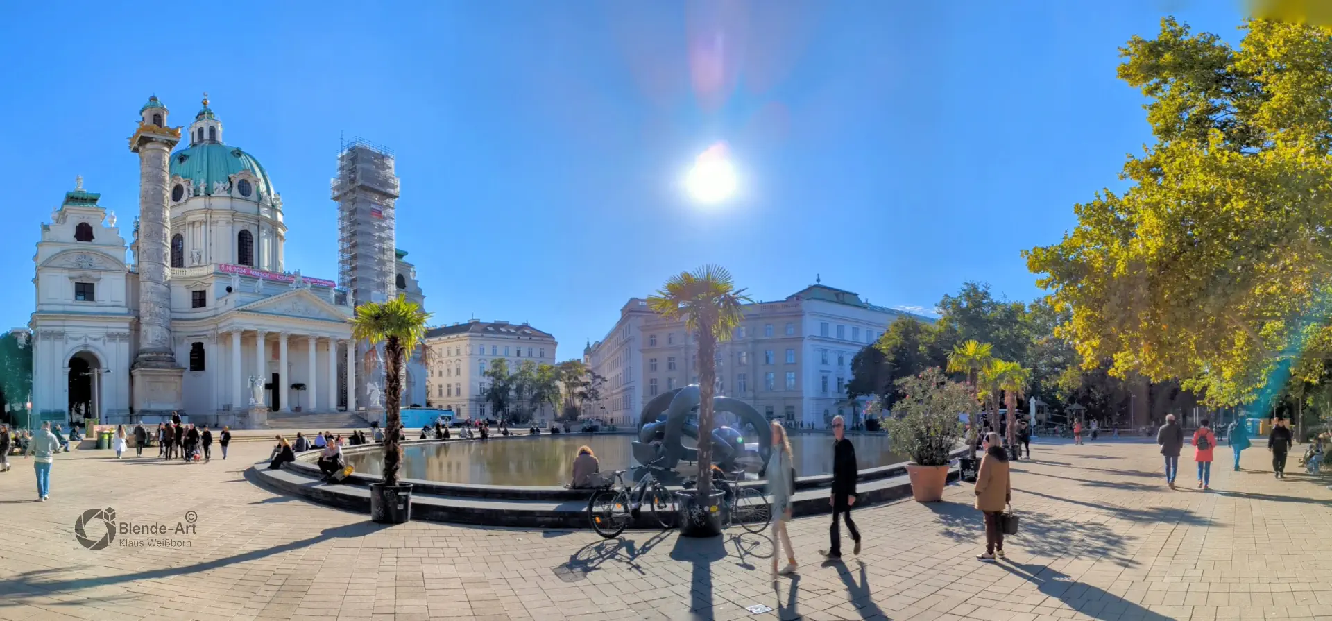 Panorama-Ansicht des Wiener Karlsplatzes mit der barocken Karlskirche, dem vorgelagerten Brunnen und herbstlichen Bäumen unter sonnig blauem Himmel.