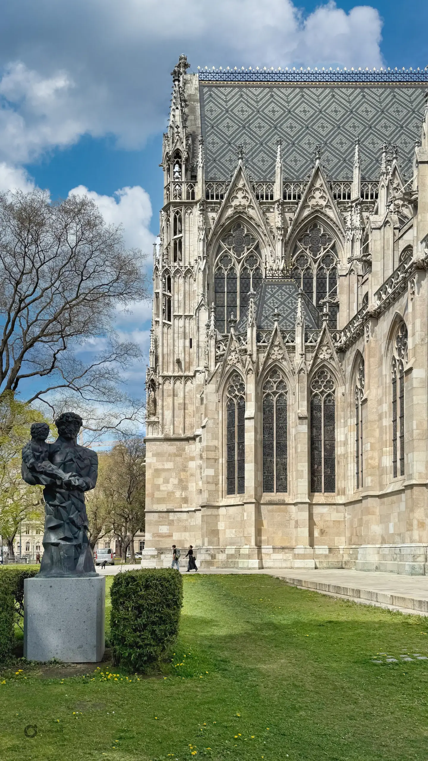 Detailansicht der neugotischen Fassade der Wiener Votivkirche mit verzierten Spitzbögen, Strebewerk und gemustertem Dach unter blauem Himmel.