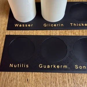 Zwei weiße 3D-gedruckte Messbecher auf beschriftetem Organisations-Board mit Markierungen für Wasser, Glycerin, Nutilis und Guarkernmehl