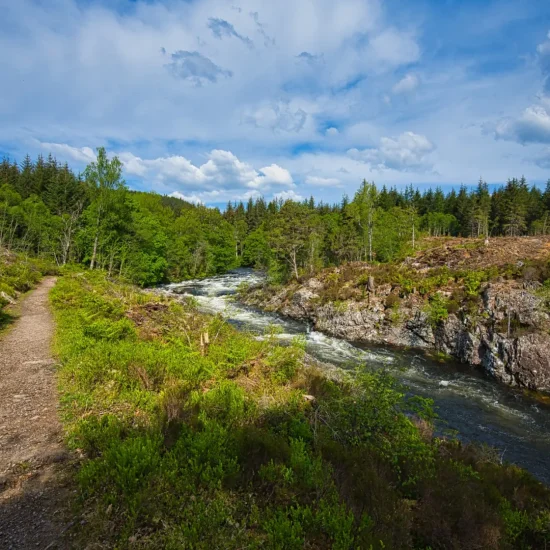 Schmaler Wanderpfad entlang des wilden River Oich mit bewaldeter Landschaft und Felsufern in den Highlands.