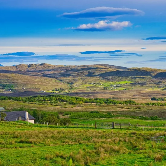 Weitläufige grüne Hügellandschaft mit vereinzelten Häusern unter einem wolkigen blauen Himmel auf der Isle of Skye.