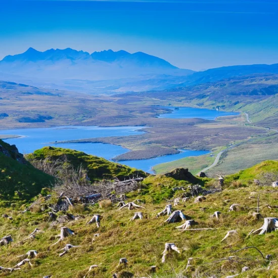 Panoramablick von einem Hügel auf der Isle of Skye über mehrere Lochs und weitläufige Hügel, mit den Cuillin Hills in der Ferne. Baumstümpfe im Vordergrund und eine kurvige Straße führen durch die Landschaft.