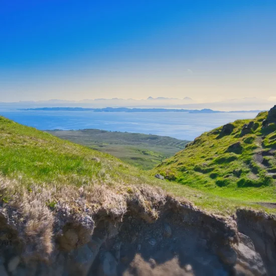 Weitläufiger Blick vom grünen Hügelpfad der Isle of Skye hinüber zur schottischen Küste, mit leuchtend blauem Himmel und dem Meer in der Ferne.
