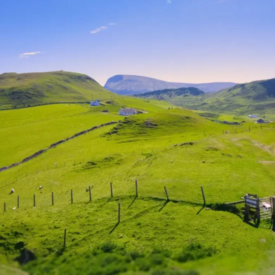 Panorama mit grünem Weideland und vereinzelten weißen Häusern vor Hügeln und Bergen bei Staffin auf der Isle of Skye, aufgenommen an einem sonnigen Tag.