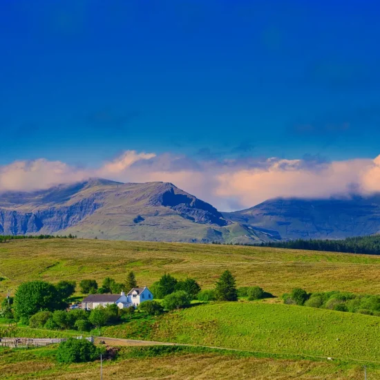 Weißes Cottage in grüner Hügellandschaft auf der Isle of Skye mit Blick auf das Storr-Massiv, dessen Gipfel teils von Wolken verhüllt sind.