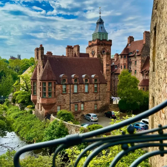 Blick auf Dean Village in Edinburgh mit roten Sandsteinhäusern, Uhrturm und Flusslauf bei Sonnenschein.