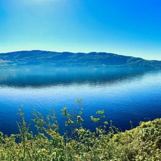 Panoramablick über Loch Ness mit glattem Wasser, grünen Uferhügeln und strahlend blauem Himmel, aufgenommen bei Urquhart Castle.