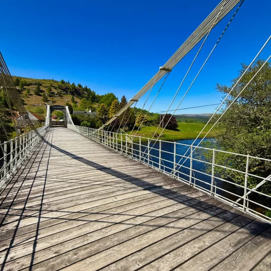 Historische Hängebrücke Bridge of Oich mit Holzboden und Tragseilen über dem River Oich bei blauem Himmel.