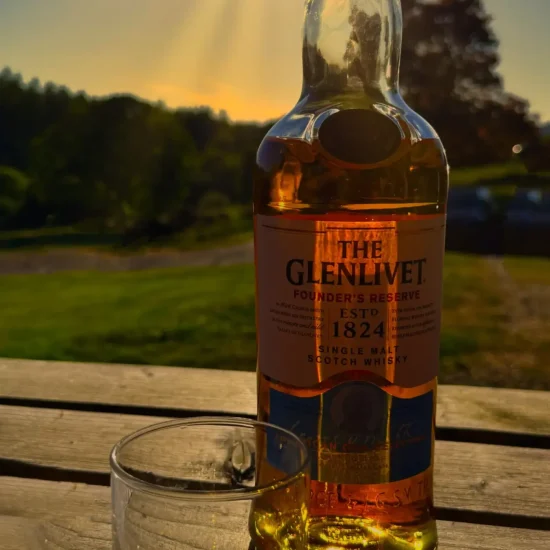 Whiskyflasche „The Glenlivet“ mit Glas im Sonnenuntergang auf Holzbank, warme Lichtstimmung.