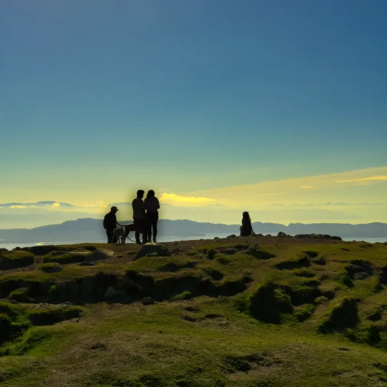 Menschen und ein Hund stehen in der Abendsonne auf einem Hügel nahe dem Old Man of Storr mit Blick auf das Meer und ferne Inseln