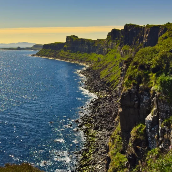 Steile grüne Klippen an der Küste der Isle of Skye, die in das tiefblaue Meer abfallen. Sonnenlicht glitzert auf der Wasseroberfläche, mit dramatischer Felskante im Vordergrund.