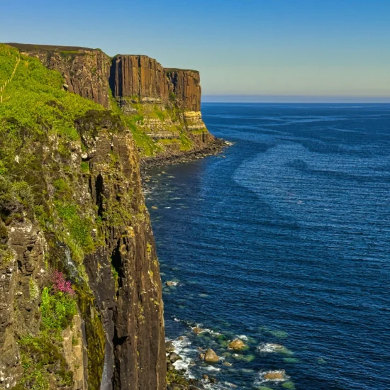 Hohe Basaltklippen mit grünem Bewuchs fallen steil ins blaue Meer ab. Rechts der Blick in die Weite des Atlantiks, links ein kleiner Wasserfall über der Felskante.