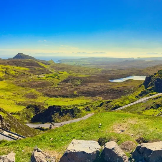 Panoramablick vom Quiraing mit Serpentinenstraße im Vordergrund, grünen Hügeln, Felsen und weiter Aussicht bis zur Küste unter klarem Himmel.